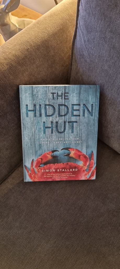 A picture of the Hidden Hut cook book on our sofa at home, ready for use. The book features countless recipes from the cafe, as well as stories of the venue. The cover features a crab claw on a blue wooden board background. The author is the Hidden Hut's owner Simon Stallard. 
